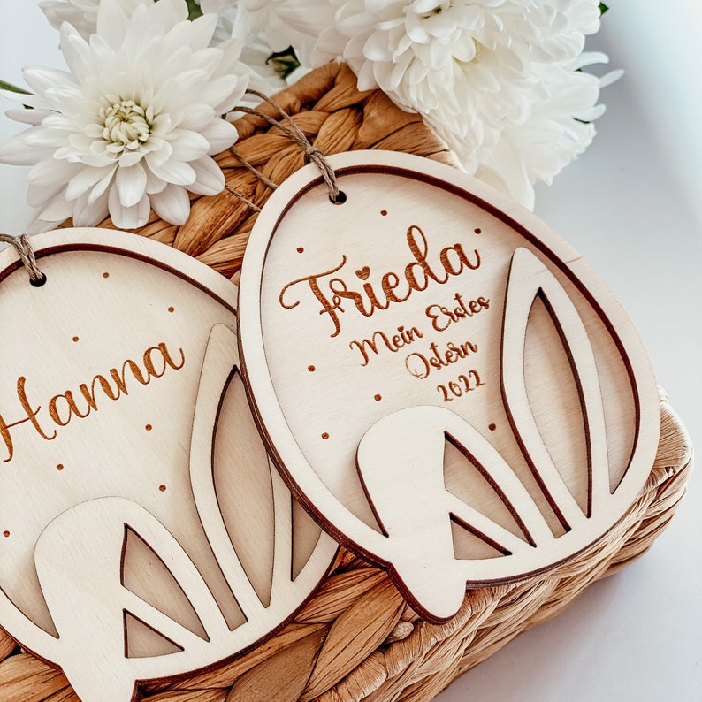 May include: Two wooden Easter egg ornaments with bunny cutouts. One ornament says "Hanna" and the other says "Frieda. My first Easter 2022".