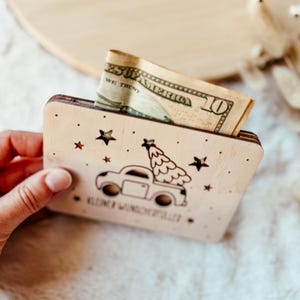 K&ouml;nnte beinhalten: Ein h&ouml;lzerner Geldhalter mit einem Design eines Trucks mit Weihnachtsbaum. Er hat Sterne und den Text "Kleine Wunderfuller". Eine Zehn-Dollar-Note ist teilweise sichtbar.