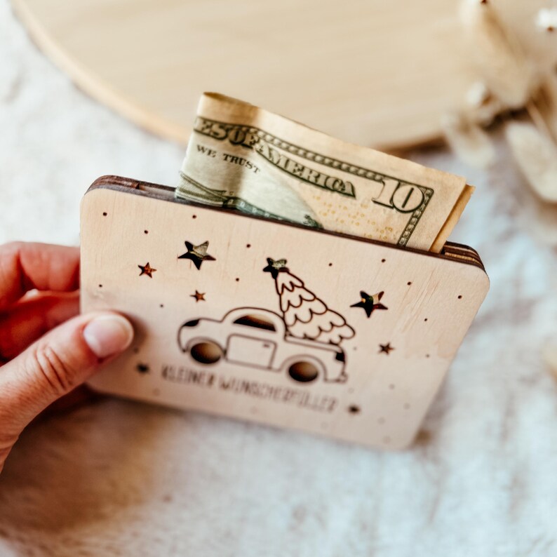 K&ouml;nnte beinhalten: Ein h&ouml;lzerner Geldhalter mit einem Design eines Trucks mit Weihnachtsbaum. Er hat Sterne und den Text "Kleine Wunderfuller". Eine Zehn-Dollar-Note ist teilweise sichtbar.