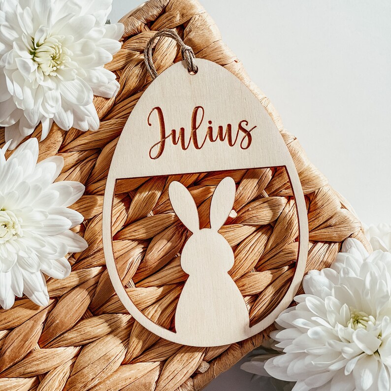 Op de afbeelding: Een houten paasei-versiering met de naam "Julius" erop gegraveerd. De versiering is versierd met een uitgesneden konijn en ligt op een geweven rieten mand.