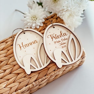 May include: Two wooden Easter egg ornaments with bunny ears. One ornament says "Hanna" and the other says "Frieda. My first Easter 2022".