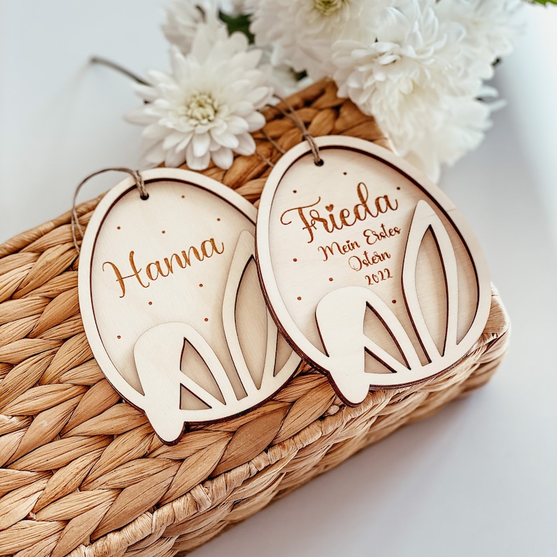 May include: Two wooden Easter egg ornaments with bunny ears. One ornament says "Hanna" and the other says "Frieda. My first Easter 2022".