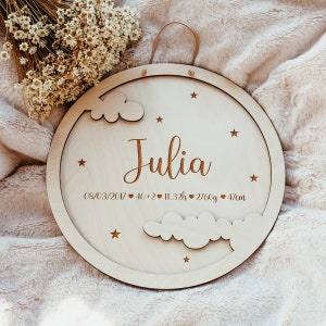 May include: A round wooden sign with a white background and a cutout design of clouds and stars. The sign says "Julia" in cursive script and includes the date "08/03/2017", followed by heart symbols and the baby's birth statistics: 40+2, 11.32h, 2760g, 47cm.