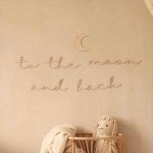 May include: Wooden wall decor featuring the phrase "to the moon and back" in cursive script. A small wooden crescent moon is positioned above the text. A stuffed bunny rabbit is in a wicker basket below the decor.
