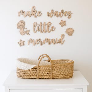 Może przedstawiać: Tkany wiklinowy kosz z białym wnętrzem stoi na białej powierzchni. Powyżej, drewniane wycięcia z napisem "make waves little mermaid" z kształtami syreny, rozgwiazdy i muszli. Dekoracja pokoju dziecięcego.