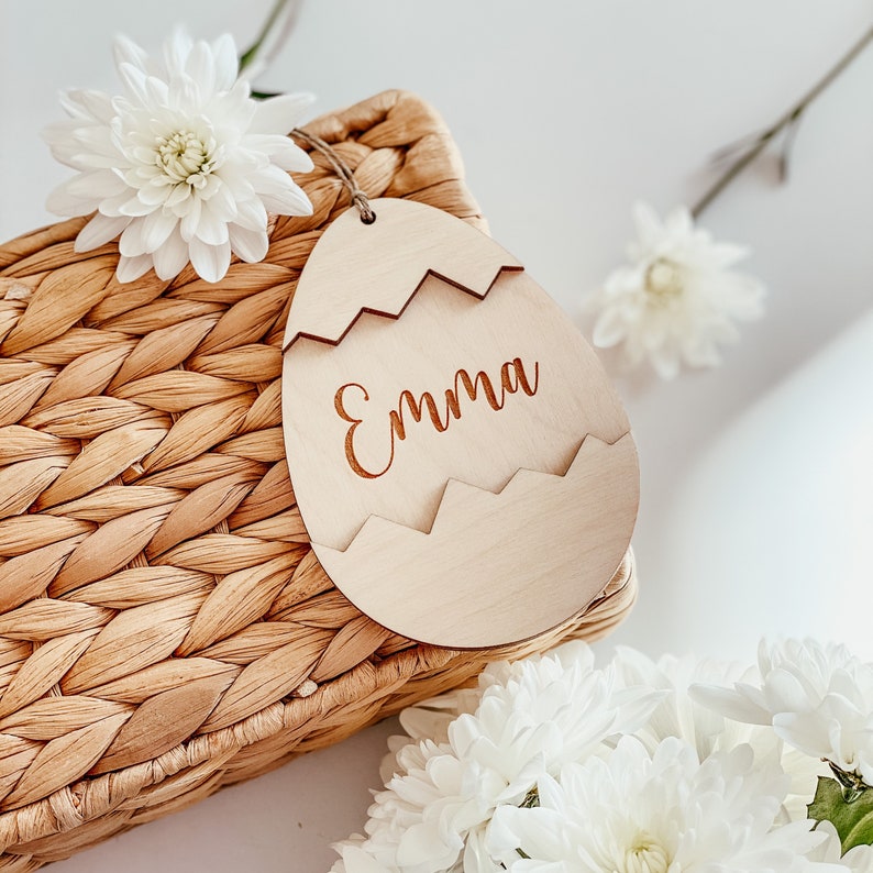 May include: A wooden Easter egg-shaped tag with the name "Emma" engraved on it. The tag is sitting on a woven basket with white flowers.