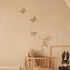 Pu&ograve; includere: Decorazioni murali in legno a forma di farfalle e fata in una cameretta. Le farfalle sono disposte in diagonale, con una sagoma di fata sottostante. Sono visibili anche una culla intrecciata e un peluche a forma di coniglietto.