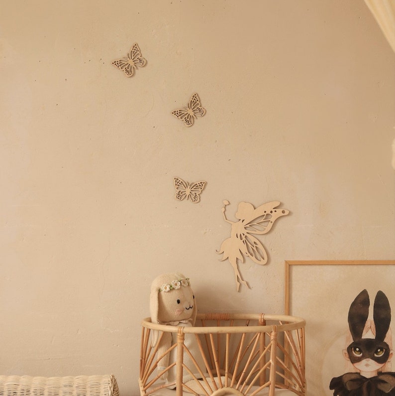 Pu&ograve; includere: Decorazioni murali in legno a forma di farfalle e fata in una cameretta. Le farfalle sono disposte in diagonale, con una sagoma di fata sottostante. Sono visibili anche una culla intrecciata e un peluche a forma di coniglietto.