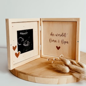 May include: A wooden keepsake box, open to reveal an ultrasound image and the text "Ihr werdet Oma & Opa". The box features two wooden heart cutouts and a small heart symbol. The box is displayed on a wooden surface.