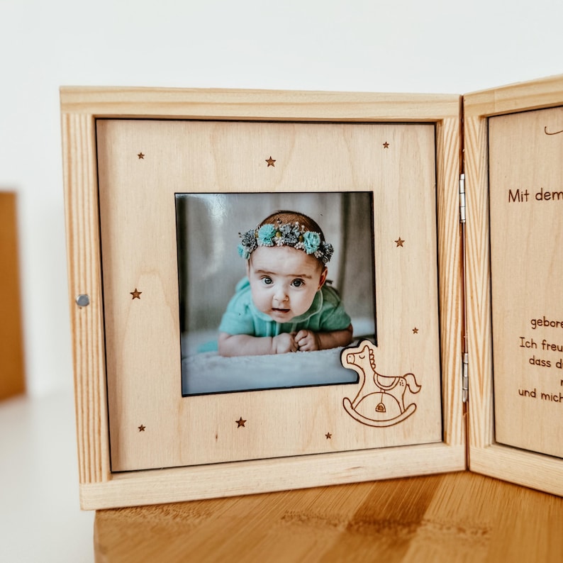 May include: A wooden photo frame with a picture of a baby wearing a flower crown. The frame has a rocking horse design and the text "Mit dem" is visible on the right side.