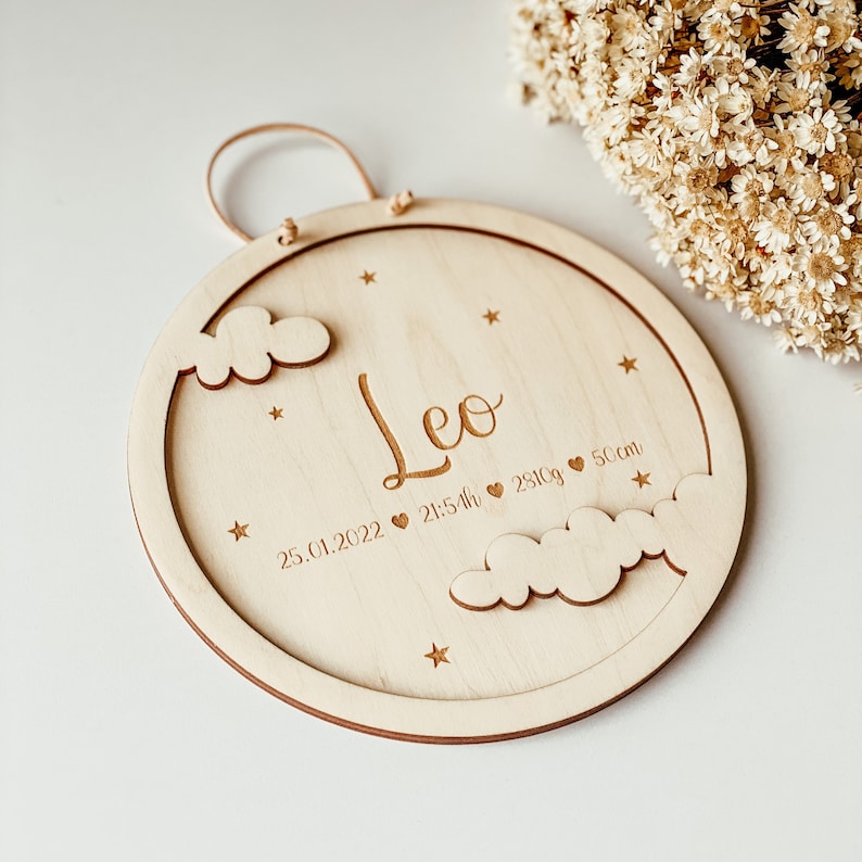 May include: A round wooden plaque with a cutout of clouds and stars. The plaque is engraved with the name "Leo", the date "25.01.2022", the time "21:54h", the weight "2810g", and the length "50cm".