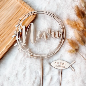 May include: Wooden cake topper with a circle wreath and the name "Nevio" in cursive. A separate wooden fish-shaped topper says "Zur Taufe 15.05.2022".