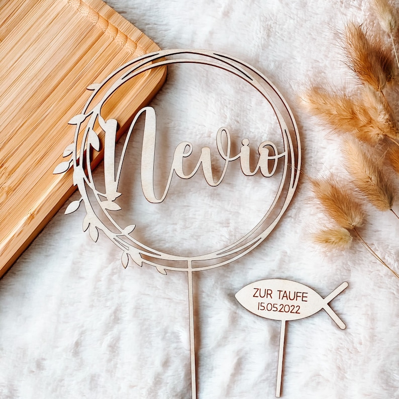 May include: Wooden cake topper with a circle wreath and the name "Nevio" in cursive. A separate wooden fish-shaped topper says "Zur Taufe 15.05.2022".