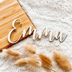 Puede incluir: Un letrero de nombre de madera con el nombre "Emma" escrito en cursiva. El letrero est&aacute; sobre una superficie blanca y esponjosa con hierba de la pampa seca.