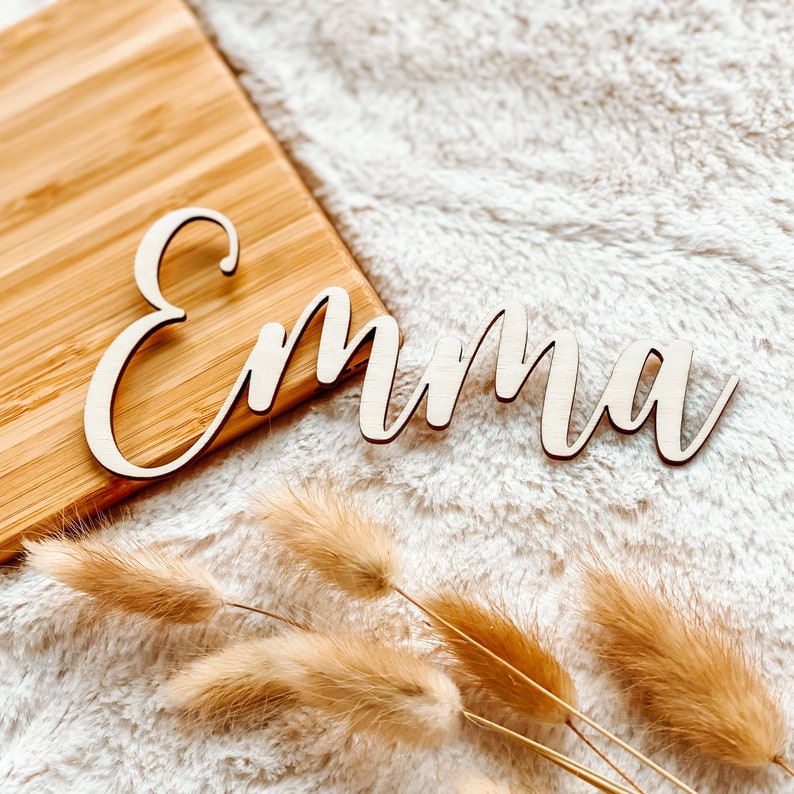 Puede incluir: Un letrero de nombre de madera con el nombre "Emma" escrito en cursiva. El letrero est&aacute; sobre una superficie blanca y esponjosa con hierba de la pampa seca.