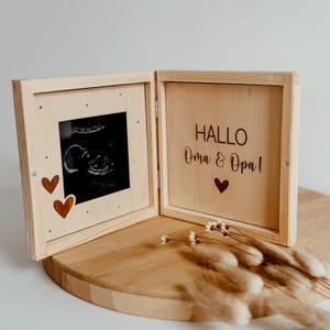 May include: A light-coloured wooden box with an ultrasound image and the text "HALLO Oma & Opa!" inside. The box has a hinged design, with a small heart detail and two wooden hearts on the left side. The box is open, revealing the contents.