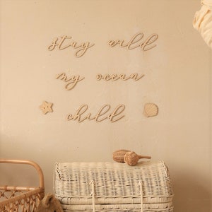 Op de afbeelding: Houten wanddecoratie met de woorden "Stay wild my ocean child" in een cursief schrift. De decoratie is getoond op een lichtbeige muur met een witte rieten kist en houten speelgoed op de voorgrond.