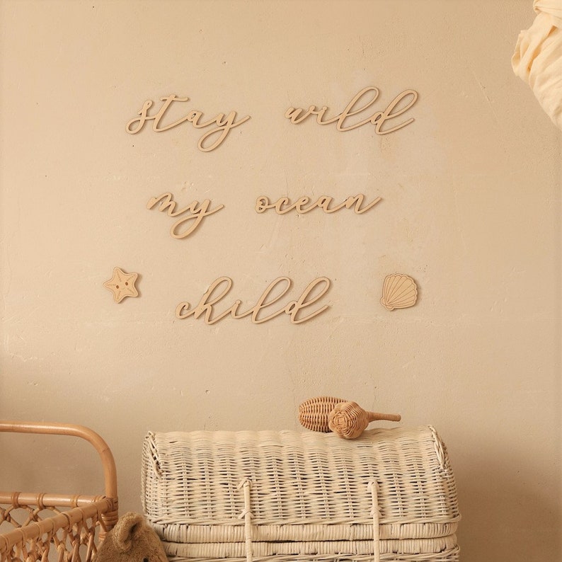 Op de afbeelding: Houten wanddecoratie met de woorden "Stay wild my ocean child" in een cursief schrift. De decoratie is getoond op een lichtbeige muur met een witte rieten kist en houten speelgoed op de voorgrond.