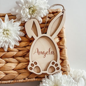 Pu&ograve; includere: Taglio di coniglio pasquale in legno con il nome personalizzato "Angela" inciso sul davanti. Il coniglio &egrave; seduto su un cesto intrecciato con fiori bianchi.