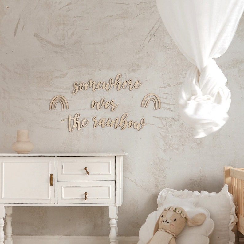 K&ouml;nnte beinhalten: Wanddekoration aus Holz mit dem Spruch "somewhere over the rainbow" und zwei Regenbogen-Ausschnitten. Die Dekoration ist an einer hellgrauen Wand in einem Kinderzimmer angebracht.