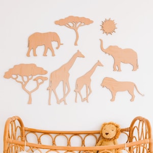 May include: Wooden safari animal wall decor set featuring an elephant, giraffe, lion, trees, and sun. The light brown cutouts are arranged on a white wall above a wicker crib with a stuffed lion toy.