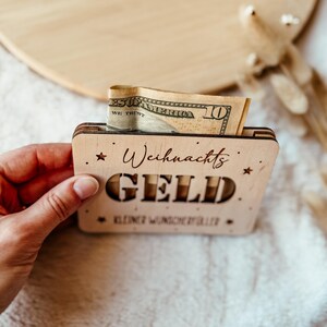 May include: A small, wooden box with the words "Weihnachts GELD" cut out, holding a ten-dollar bill. The box is decorated with star shapes and additional text. The box is held by a person.