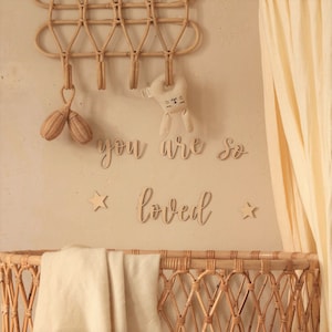 Op de afbeelding: Een wieg van gevlochten wilg met een wit dekentje en een houten bord met de tekst "you are so loved" die aan de muur erboven hangt. Er hangen twee houten sterren en een houten konijnenknuffel aan de muur.