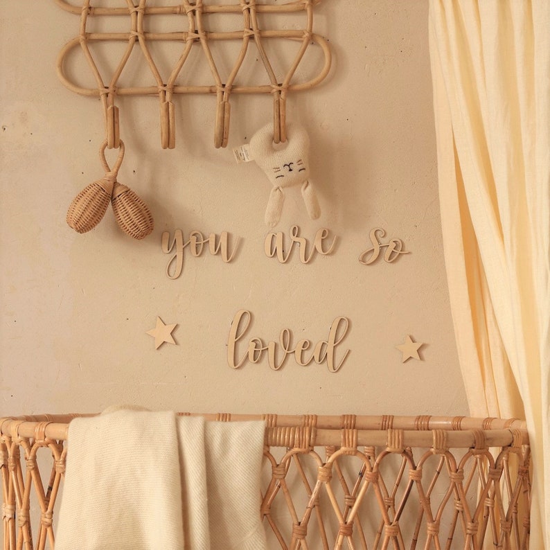 May include: A wicker crib with a white blanket and a wooden sign that reads "you are so loved" hanging on the wall above it. There are two wooden stars and a wooden bunny toy hanging from the wall.