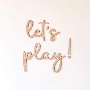 May include: Wooden wall art spelling out "let's play!" in a cursive font. The letters are a light brown color and are mounted on a white wall. The exclamation point is a separate piece.