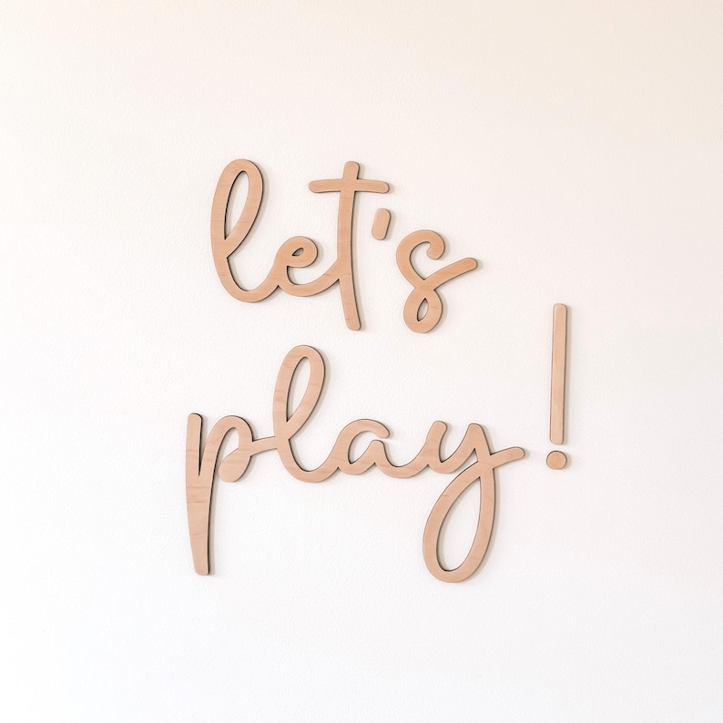 May include: Wooden wall art spelling out "let's play!" in a cursive font. The letters are a light brown color and are mounted on a white wall. The exclamation point is a separate piece.