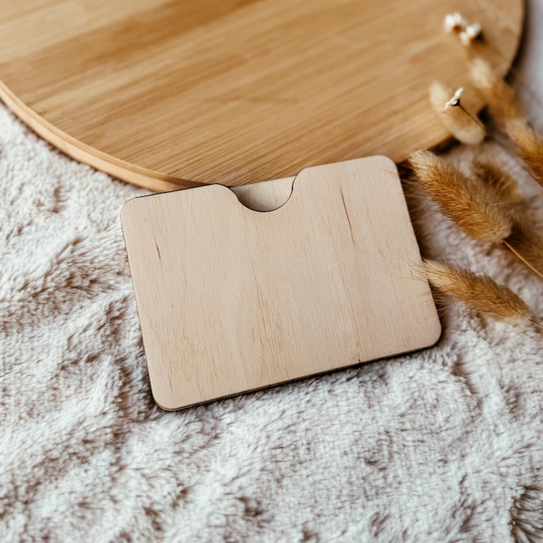 May include: A light-colored wooden card holder with a curved top cutout, resting on a soft, white, textured surface. A round wooden tray is in the background. Dried wheat stalks are to the right.