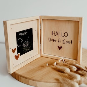 May include: A wooden keepsake box, hinged to open, displaying an ultrasound scan on the left and the text "HALLO Oma & Opa!" with a heart on the right. The box is open, revealing the contents. Decorative hearts are on the left panel.