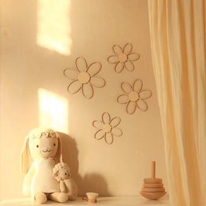 Peut inclure: Un lapin en peluche beige tricoté avec une couronne de fleurs est assis sur une surface blanche. La poupée tient un plus petit animal en peluche beige tricoté. L'arrière-plan présente un mur beige avec cinq décorations murales en bois en forme de marguerite.