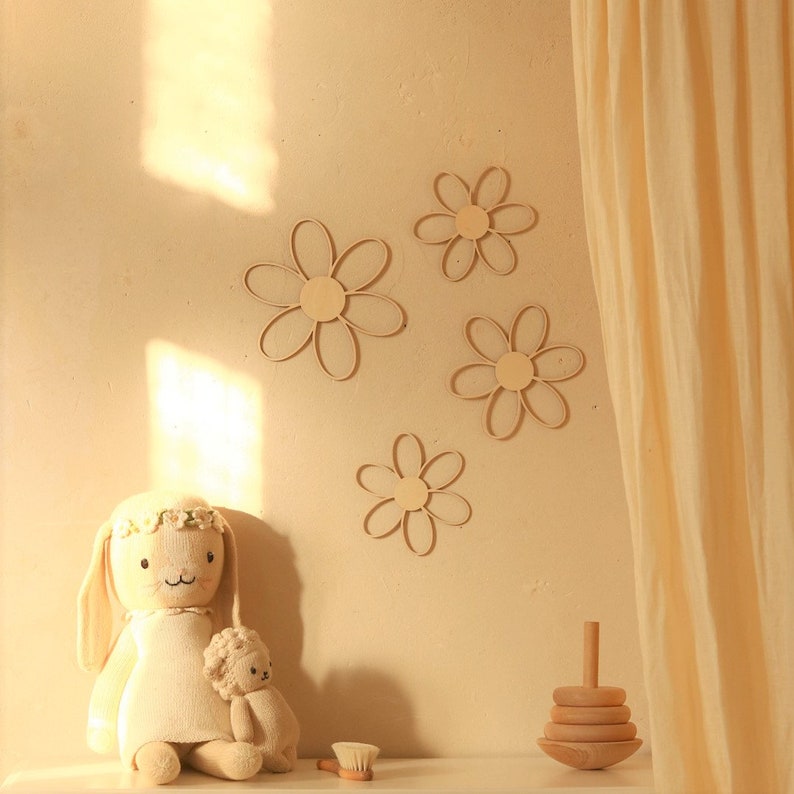 May include: A beige knitted bunny doll with a flower crown sits on a white surface. The doll is holding a smaller beige knitted animal. The background features a beige wall with five wooden daisy-shaped wall decorations.