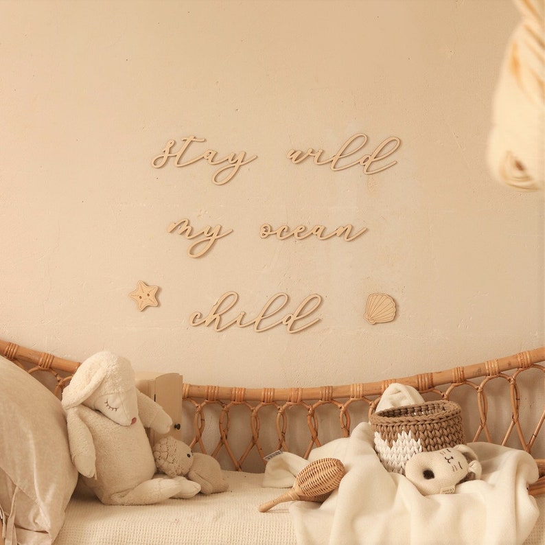 Op de afbeelding: Houten wanddecoratie met de woorden "Stay wild my ocean child" in een cursief schrift. De decoratie is bevestigd aan een lichtbeige muur boven een wieg van gevlochten riet met een wit dekbed en knuffels.