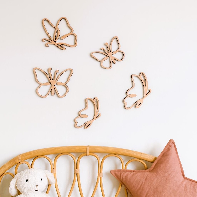 May include: Five wooden butterfly wall decorations in various designs are displayed against a white wall. The butterflies are light brown and appear to be cut from wood. A rattan headboard, a stuffed animal, and a star-shaped pillow are visible at the bottom.