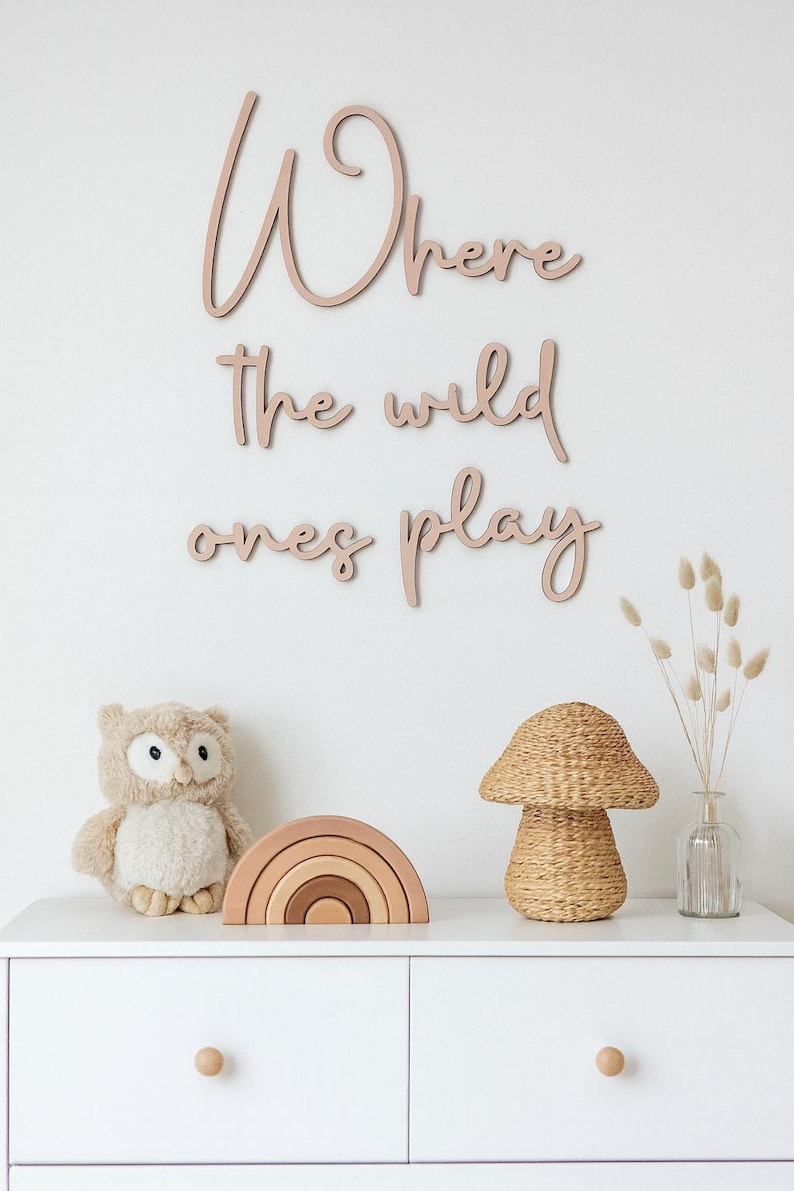 May include: A white chest of drawers with a wooden sign saying "Where the wild ones play." It's decorated with a stuffed owl, a wooden rainbow, a woven mushroom lamp, and a vase with dried flowers.