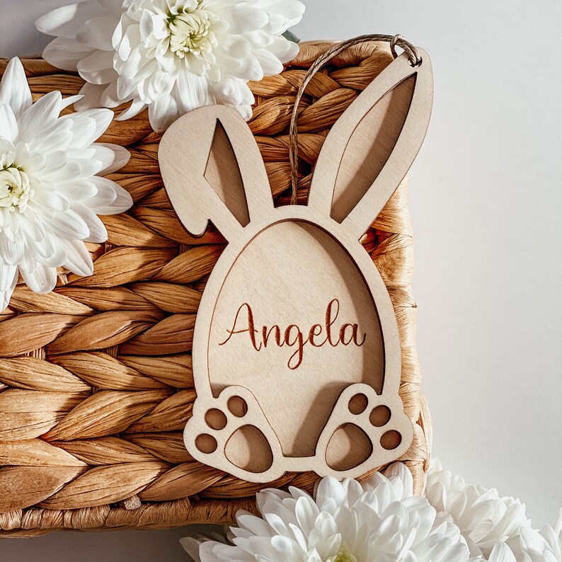 Pu&ograve; includere: Porta uova di Pasqua in legno a forma di coniglio con il nome "Angela" inciso sul davanti. Il porta uova &egrave; appoggiato su un cesto intrecciato con fiori bianchi.