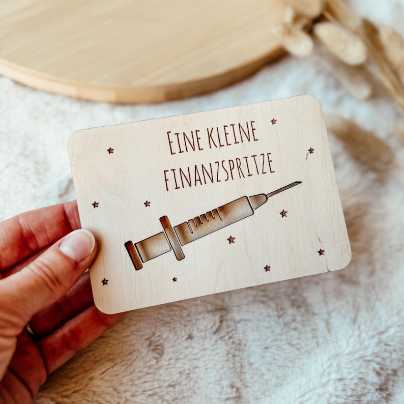 May include: A rectangular wooden plaque with the German text "EINE KLEINE FINANZSPRITZE" and a syringe illustration. The design includes small star accents. The plaque is held by a hand.