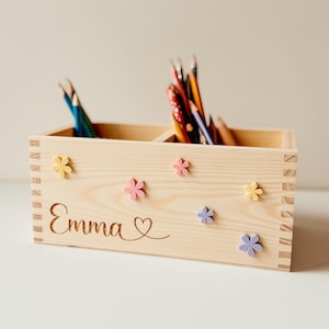 May include: A wooden pencil holder with two compartments, personalized with the name "Emma" and a heart. The holder is decorated with colorful flower-shaped embellishments.
