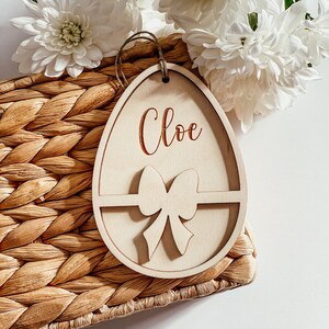 May include: A wooden Easter egg ornament with a cutout bow and the name "Chloe" engraved on the front. The ornament is sitting on a woven basket.