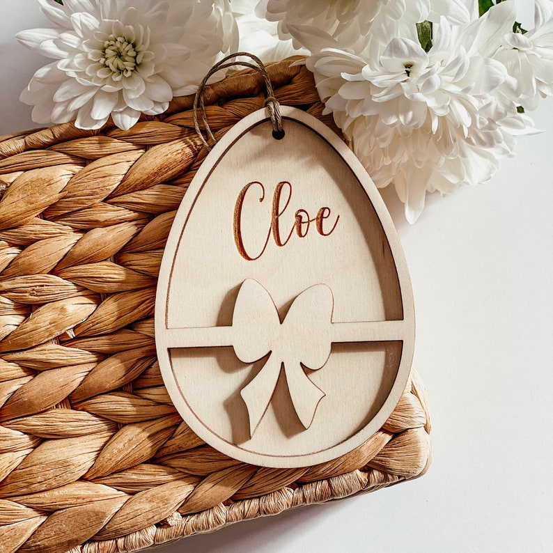 May include: A wooden Easter egg ornament with a cutout bow and the name "Chloe" engraved on the front. The ornament is sitting on a woven basket.