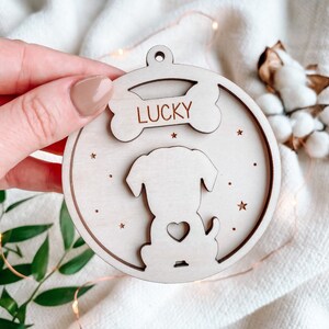 Pode incluir: Um enfeite de cachorro de madeira com um recorte em forma de osso acima da cabeça do cachorro, com a palavra "LUCKY" gravada. O cachorro tem um detalhe em forma de coração no peito. O enfeite é redondo com recortes de estrelas.