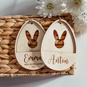 Pode incluir: Dois enfeites de ovo de P&aacute;scoa de madeira com recortes de coelho. Os enfeites s&atilde;o decorados com os nomes "Emma" e "Anton" e o texto "Mein erstes Ostern 2022".