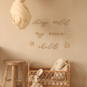 Op de afbeelding: Houten wanddecoratie met de woorden "Stay wild my ocean child" in een cursief schrift. De decoratie is bevestigd aan een beige muur met een witte stof die erboven hangt. Er is een kleine houten ster en een schelp op de muur. Een wieg van gevlochten riet met een pluche schaap en konijn staat op de voorgrond.