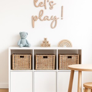 May include: A white storage unit with three woven baskets, a blue teddy bear, wooden blocks, and a rainbow-shaped toy. The wall above has the words "Let's play!" in wooden letters. A small wooden table and rug are also present.