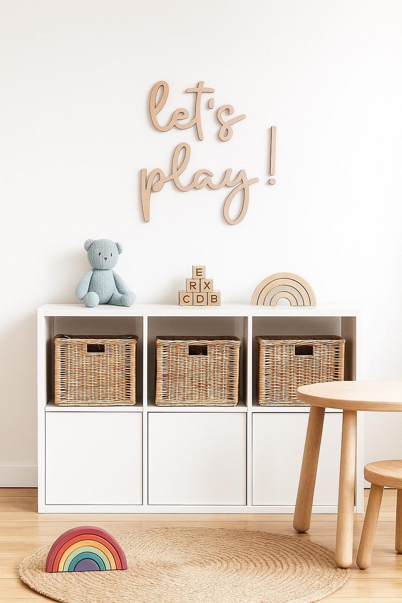 May include: A white storage unit with three woven baskets, a blue teddy bear, wooden blocks, and a rainbow-shaped toy. The wall above has the words "Let's play!" in wooden letters. A small wooden table and rug are also present.
