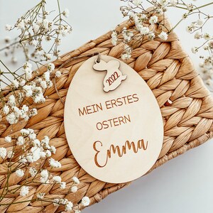 May include: A wooden Easter egg ornament with the text "Mein erstes Ostern" and the name "Emma" engraved on it. A small wooden bunny with the year 2022 is attached to the top of the egg.