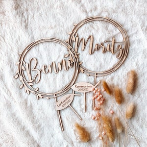 May include: Two wooden cake toppers with floral wreaths and the names "Benno" and "Moritz" inscribed. The topper for Benno has a sign that reads "Confirmation 02/01/2022" and the topper for Moritz has a sign that reads "Kommunion 01/05/2022".