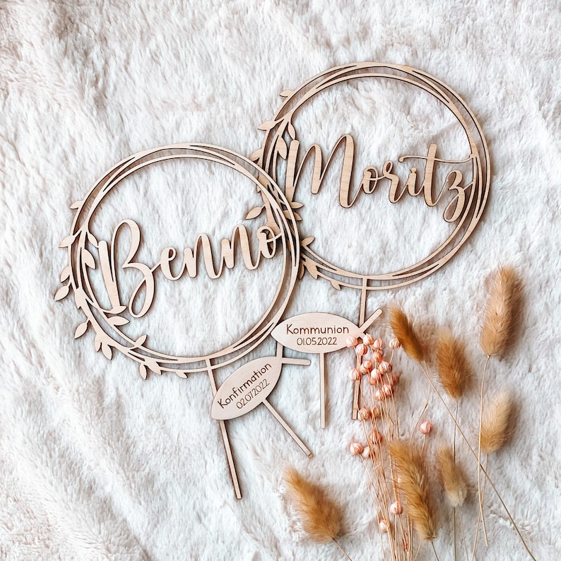 May include: Two wooden cake toppers with floral wreaths and the names "Benno" and "Moritz" inscribed. The topper for Benno has a sign that reads "Confirmation 02/01/2022" and the topper for Moritz has a sign that reads "Kommunion 01/05/2022".